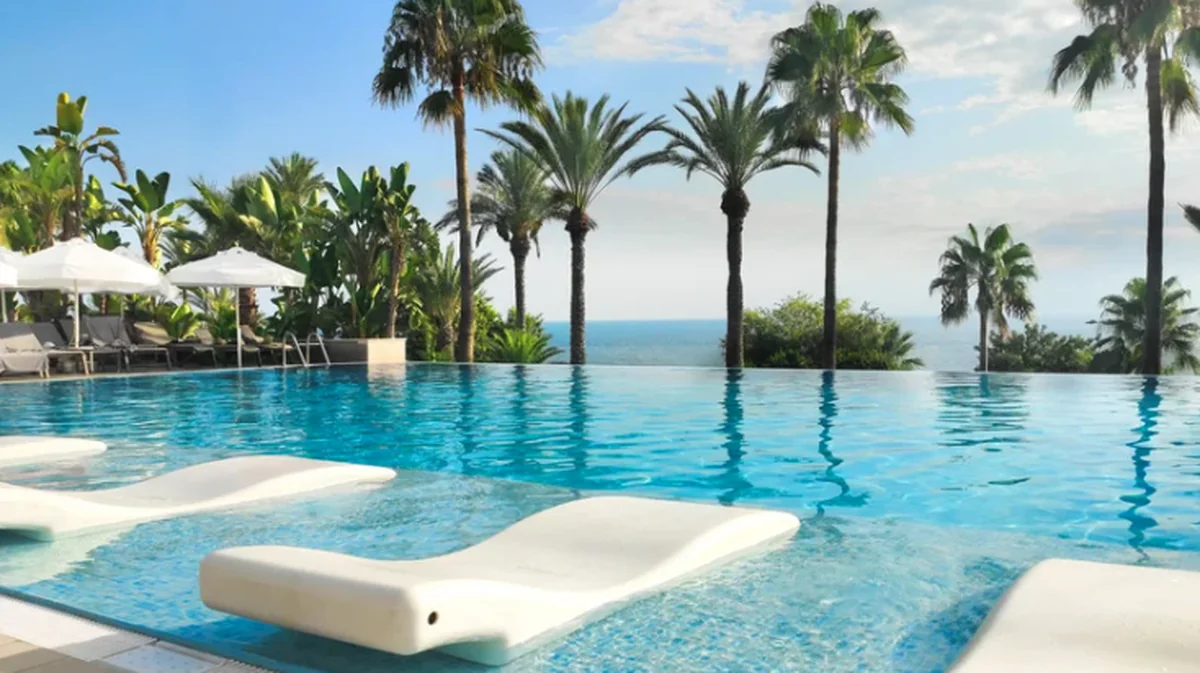An infinity pool at a luxury resort, lined with floating white loungers, palm trees framing a Mediterranean sea view beyond.