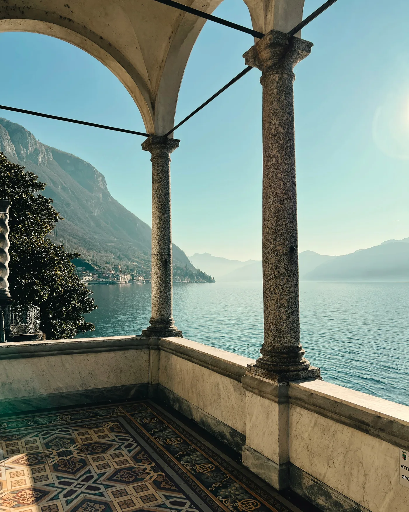 A view of Lake Como through the marble columns of a lakeside loggia, with intricate patterned floor tiles in the foreground.